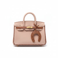 A small blush pink fuzzy faux-fur handbag with a gold-tone turn-lock clasp and a brown leather horseshoe charm, held by a hand over a cardboard box.